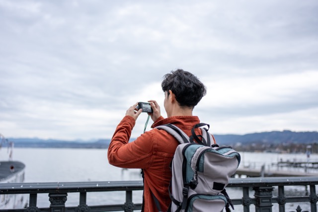 Asian backpacker use camera taking a picture of beautiful landscape. Young male tourist traveler travel alone on street in city, using cellphone record vlog on holiday vacation trip in Switzerland.